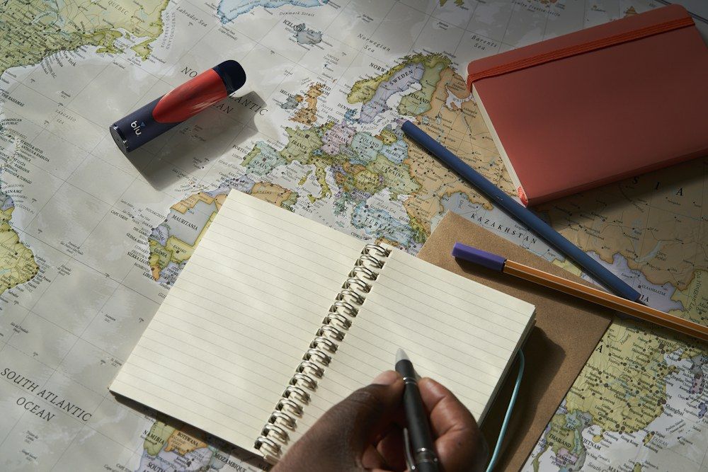 A black gel pen rests on the edge of an open spiral notebook, both placed over a colorful world map.