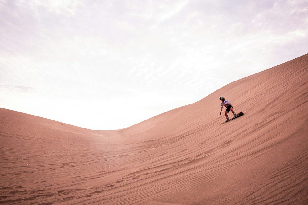 Sandboarder climbing a large reddish dune with a board