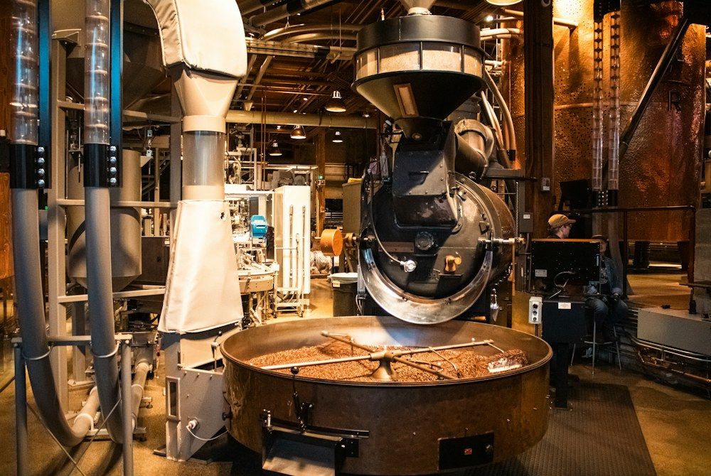 A large-scale coffee roasting machine dominates a factory floor, its drum open to release a cascade of freshly roasted beans into a circular cooling tray.