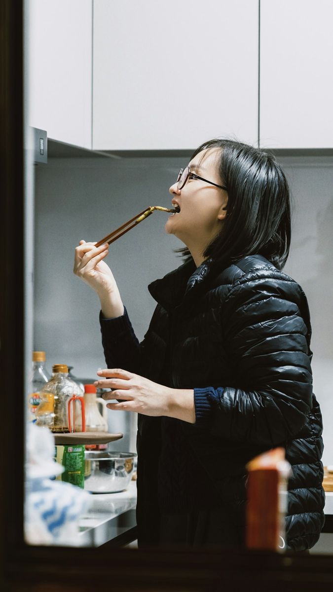 Woman in a puffer jacket tastes food from chopsticks.