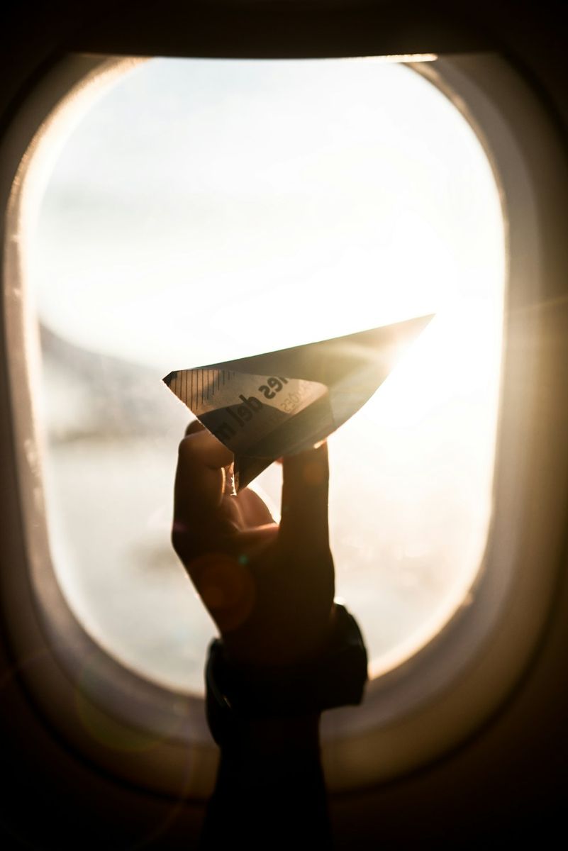 Hand holding a folded paper airplane beside a sunlit aircraft window