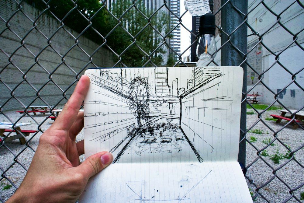 A hand holds open a sketchbook to reveal a black ink perspective drawing of a narrow alleyway, its vanishing lines converging toward a distant opening.
