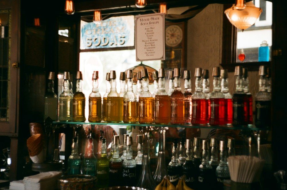 A nostalgic soda fountain counter showcases two glass shelves filled with clear bottles of house-made soda syrups.