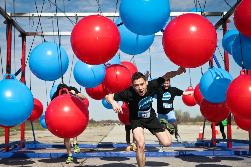 Participants in black shirts marked “Blood Sweat &” sprint beneath a series of giant red and blue hanging balls.