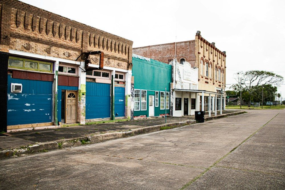 A line of aged storefronts, painted in shades of blue, green, and tan, stretches along a vacant concrete street under an overcast sky.