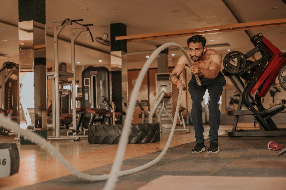A shirtless man leans forward, gripping heavy battle ropes with both hands as he performs an intense exercise.