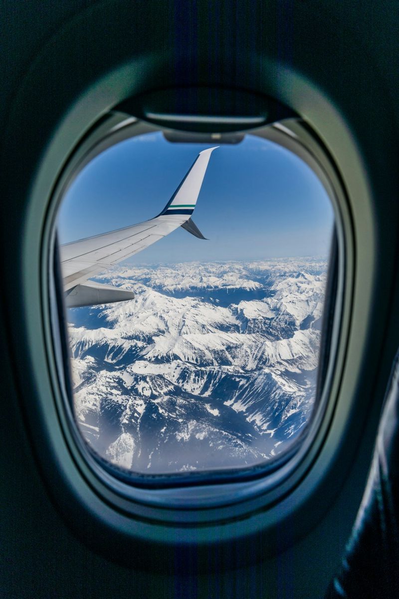 A vast expanse of snow-capped peaks stretches beneath a clear blue sky, framed by the curved edge of an airplane window.