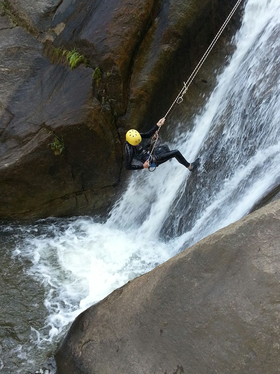 A climber equipped with a yellow helmet and black wetsuit carefully rappels down a steep, wet rock face adjacent to a roaring waterfall.